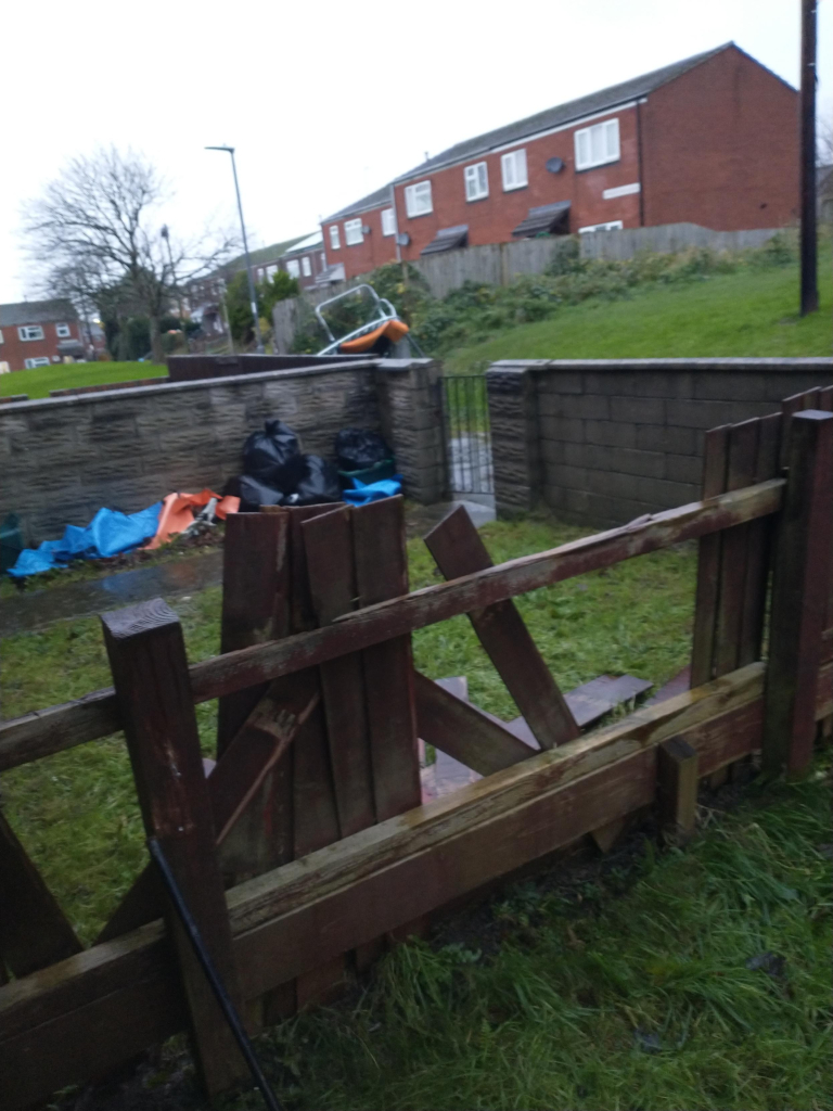 A wooden fence in the foreground missing many slats, beyond it a brick wall around another garden, and beyond that a ruined trampoline beside a path. Behind it are several other houses.