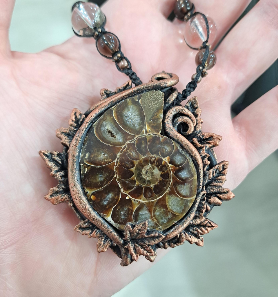 A halved and polished ammonite framed in a polymer clay design. The frame is copper colored and features swirls and small ivy leaves, matching the color scheme of the fossil. The pendant is suspended from a braided cord decorated with small quartz beads.