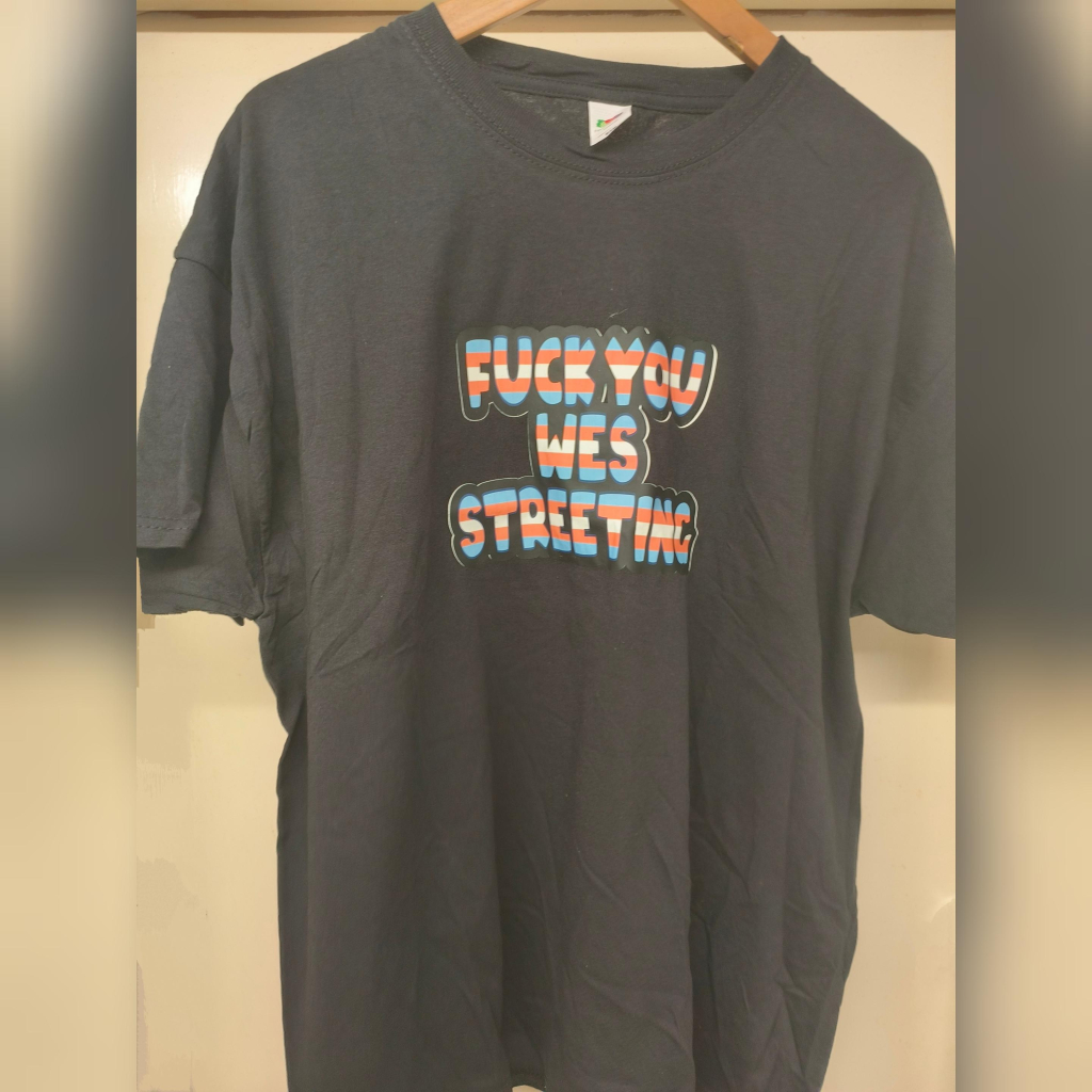 Black t-shirt with the slogan "Fuck You Wes Streeting" in the colours of the trans flag.