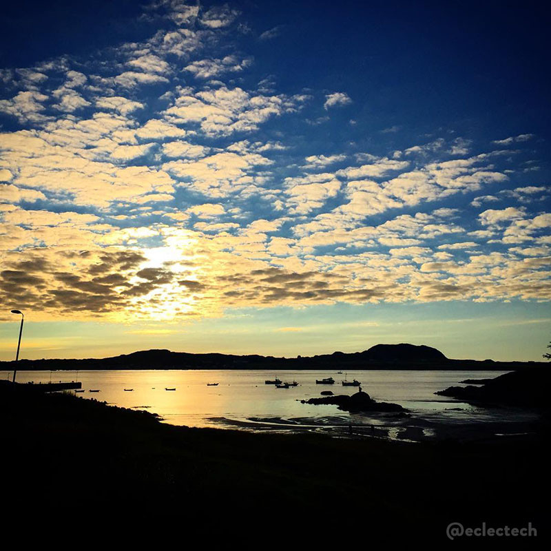 A square photo taken at sunset over the sea from the Isle of Mull to Iona. The land is a dark silhouette, the beach in the foreground and Iona a bumpy strip just below the centre. The water is golden with small boats dotted across it. The sky is blue - light near Iona and deeper blue at the top - and there are dappled clouds, dark with the bright sun behind at the bottom, fluffy and white at the top, covering the sky.