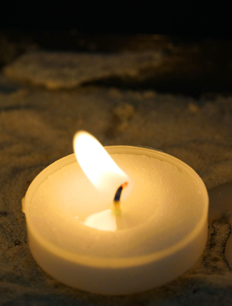 a small and short yellow tealight candle burns with a bright white-yellow flame. it's sitting in sand which is spread on a dark surface. 