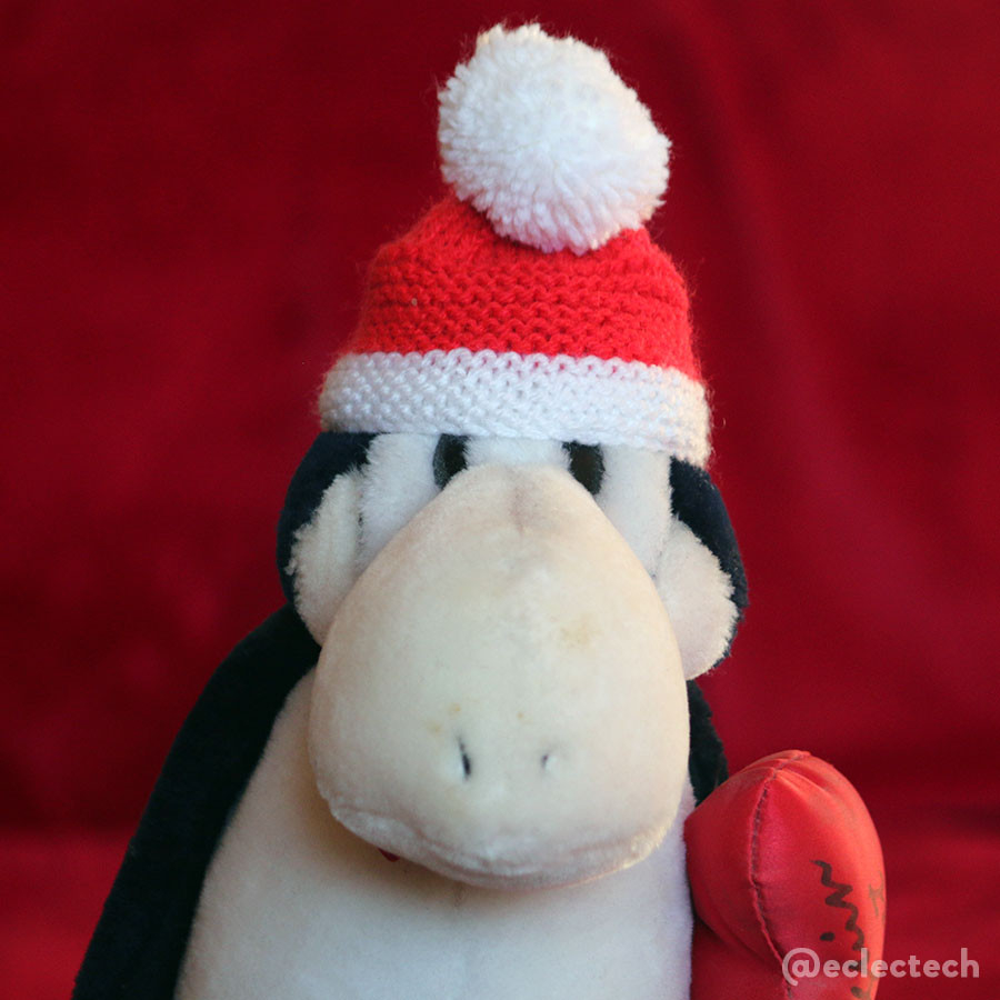 A photo of the top half of an Opus the Penguin plushie against a blurred red backdrop. He is wearing a Santa hat. 