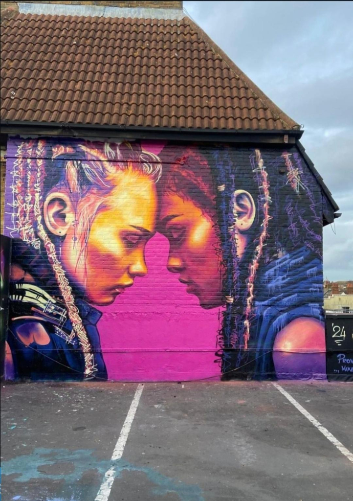 Streetartwall. A large mural showing two young women facing each other was sprayed onto a rectangular brick wall on a roof. The mural shows a young woman with long blonde dreadlocks leaning her forehead against the forehead of a young woman of color with black dreadlocks. Both have their eyes closed. The background is a vibrant pink. A really nicely done mural with a beautiful message of friendship and great colors. 
Info: Not on the photo is the artist's signature with the following text: "Shauna Anseo - Pronounced: Un-shuh - makes sense in irish."