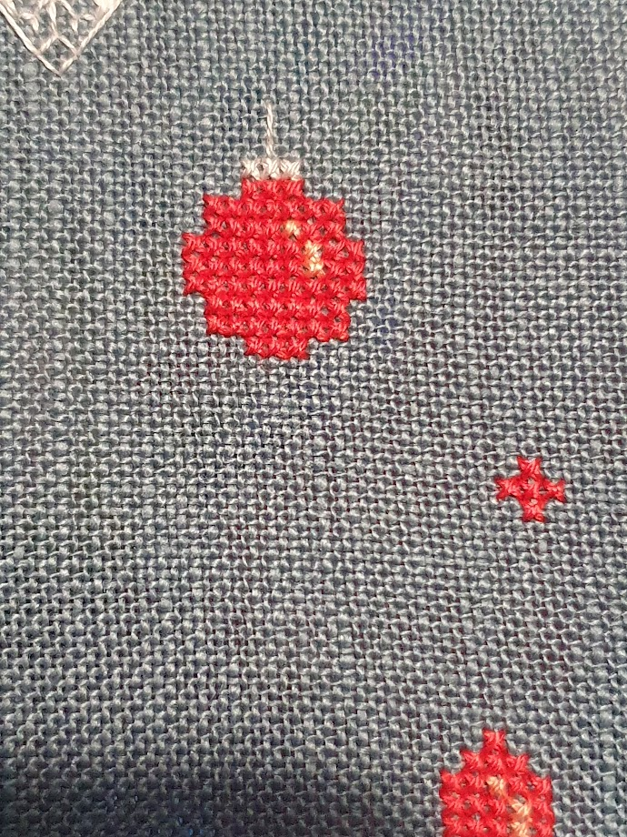 A simple round red bauble with a tin coloured top. A small red cross is  slightly lower and to the right of the red bauble. 