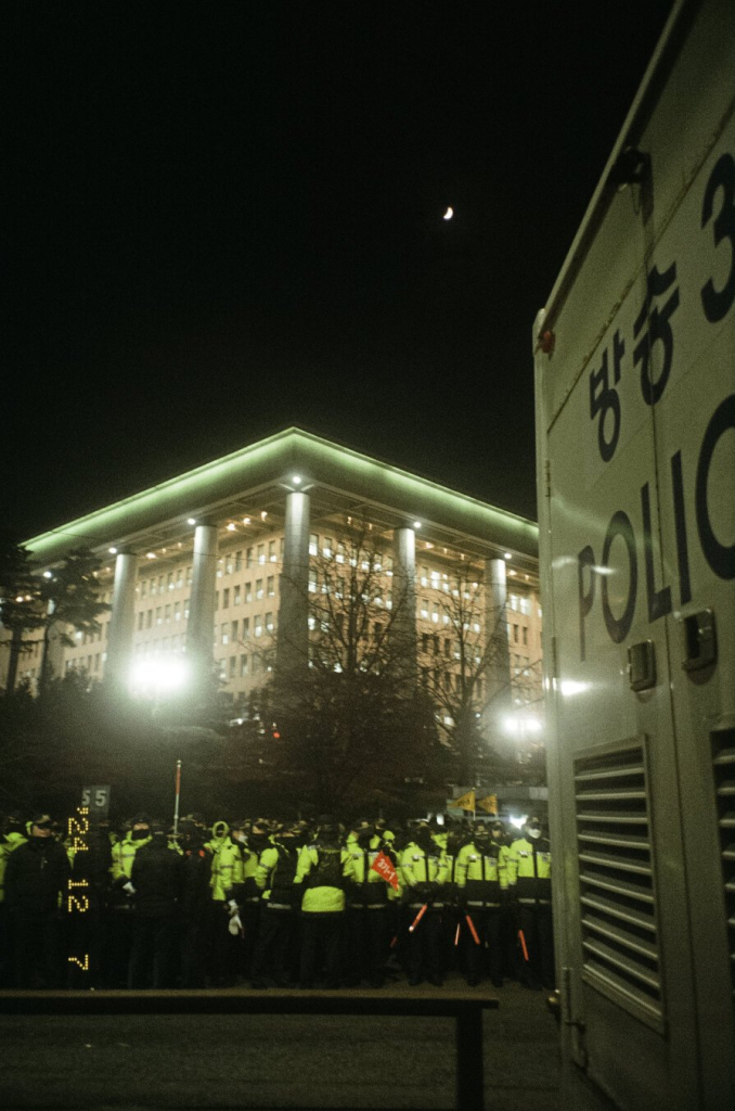 어두운 밤, 경찰 버스와 경찰 무리 뒤로 국회 건물 뒷편 모서리가 보인다. 우측 상단에는 반달이 떴다.