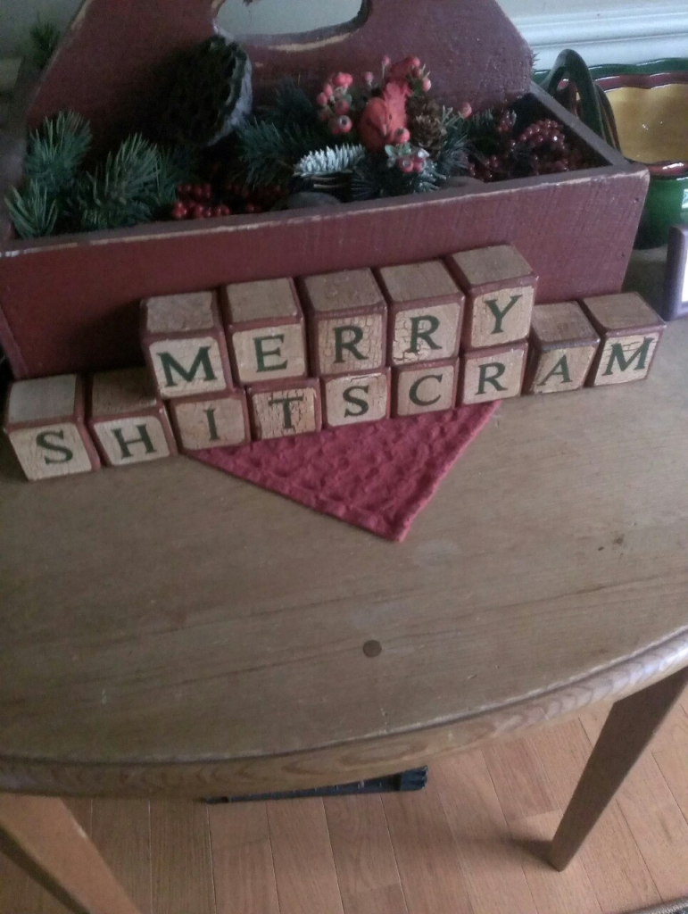 Christmas decorations on a wooden table, with wooden blocks spelling out: Merry Shitscram