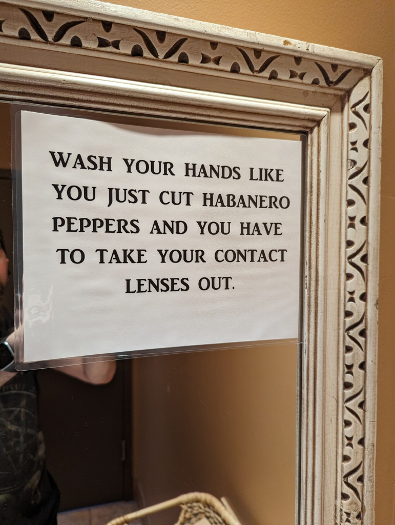 A sign affixed to the bathroom mirror that reads: "WASH YOUR HANDS LIKE YOU JUST CUT HABANERO PEPPERS AND YOU HAVE TO TAKE YOUR CONTACT LENSES OUT."