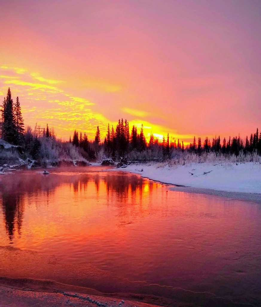 A cold winter morning, looking across a still clear river just as the sun rise begins to fill the sky with orange red hues, that also reflect on the water.