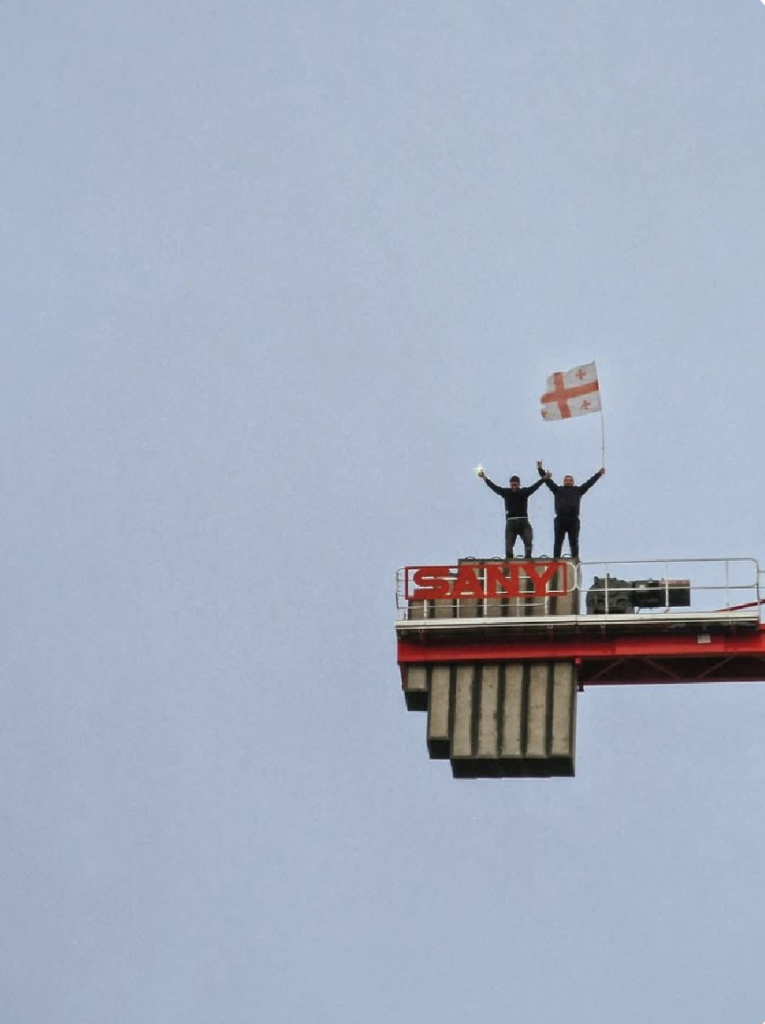 Two construction workers stand on top of a huge crane in Tbilisi, Georgia. They're waving their arms in the air and flying a huge flag of Georgia.

Photo by Dato Simonia