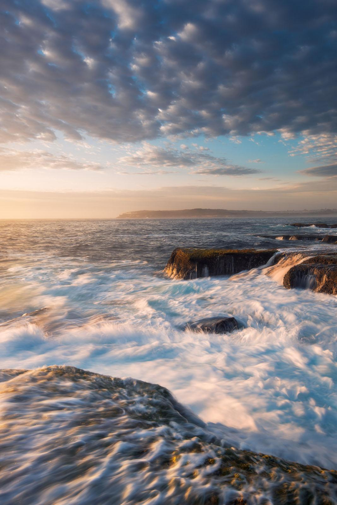 vertical seascape during sunrise. There's a rock platform in the foureground and another one in the middle. The photo catches rushing water and morning golden light