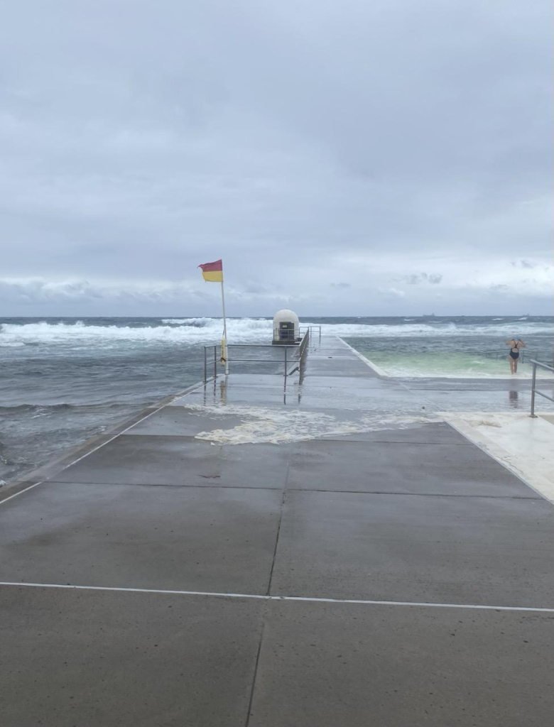 The red and yellow lifesaving flag is flying out in the strong southerly wind. It's on the concrete concourse on the northern side of the Merewether Ocean Baths. The ocean and main pools are visible on either side of the wide path. Light grey sky and darker grey water. The ocean has spilled across the path. A swimmer is starting down the entry ramp to the main pool. 