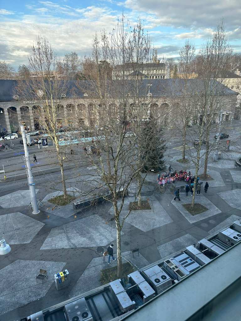 A grey winter town square, with bare trees in the middle, a train station building with high arches behind, and a group of very young children with orange tabards arranged in a choir in the middle of the square, in front of a large conifer/Christmas tree