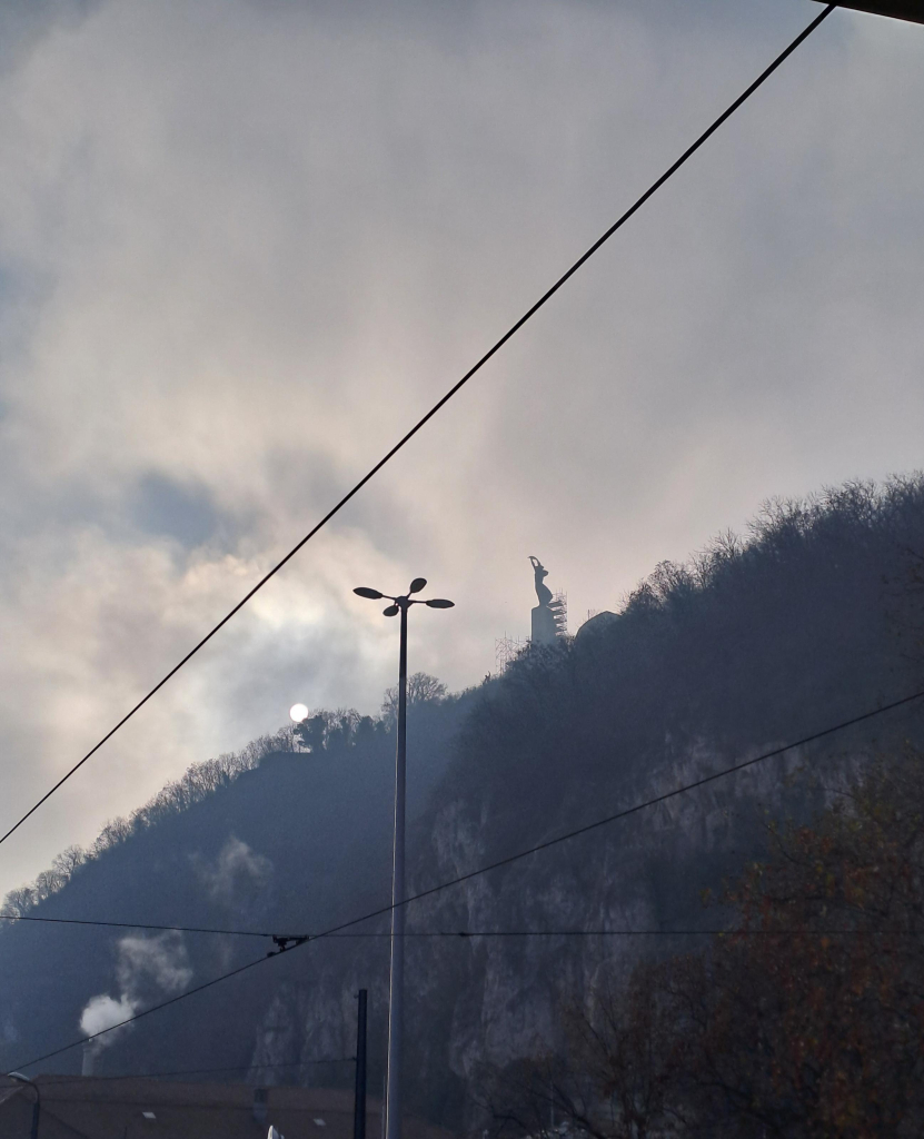 Foggy view of Gellért Hill, silhouetted against a cloudy sky. The pale disc of the sun is peeking over the tree line, surrounded by a halo of light. On top of the hill is the statue of liberty, a woman's silhouette half supported by scaffolding. Between the two, curring through the profile of the hill, is a lamppost in the foreground, branching four ways at the top. The picture is cut across by the black lines of power lines in a geometric pattern.