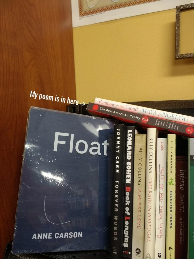 Vertical photo of an upper corner of bookshelf holding several slim volumes of poetry by Billy Collins, E. E. Cummings, Dante, and others. A volume of Anne Carson's book Float has been pulled out to reveal its simple, dark cover. Lying on top of the books are two volumes someone has left behind. Some text has been superimposed on the photo with an arrow pointing to the bottom of these two titles (Best American Poetry 2015) and reading "My poem is in here." The walls of the bookstore are warm brown wood and a sort of pale, dusky yellow.