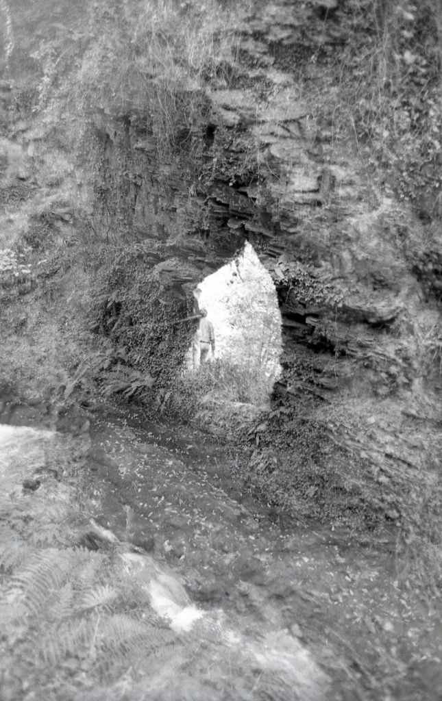 Black and white photograph. A double exposure of people hiking in the woods. In the foreground is an opening where the trail goes through a rock. The people are on the far side of the opening.