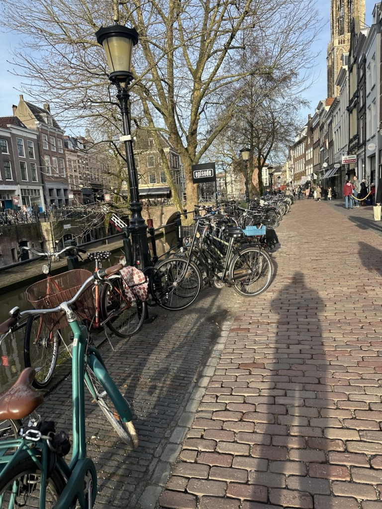 A typical Dutch canal street with a mass of parked bicycles along the side and the photographers long morning shadow stretching into the brick street foreground