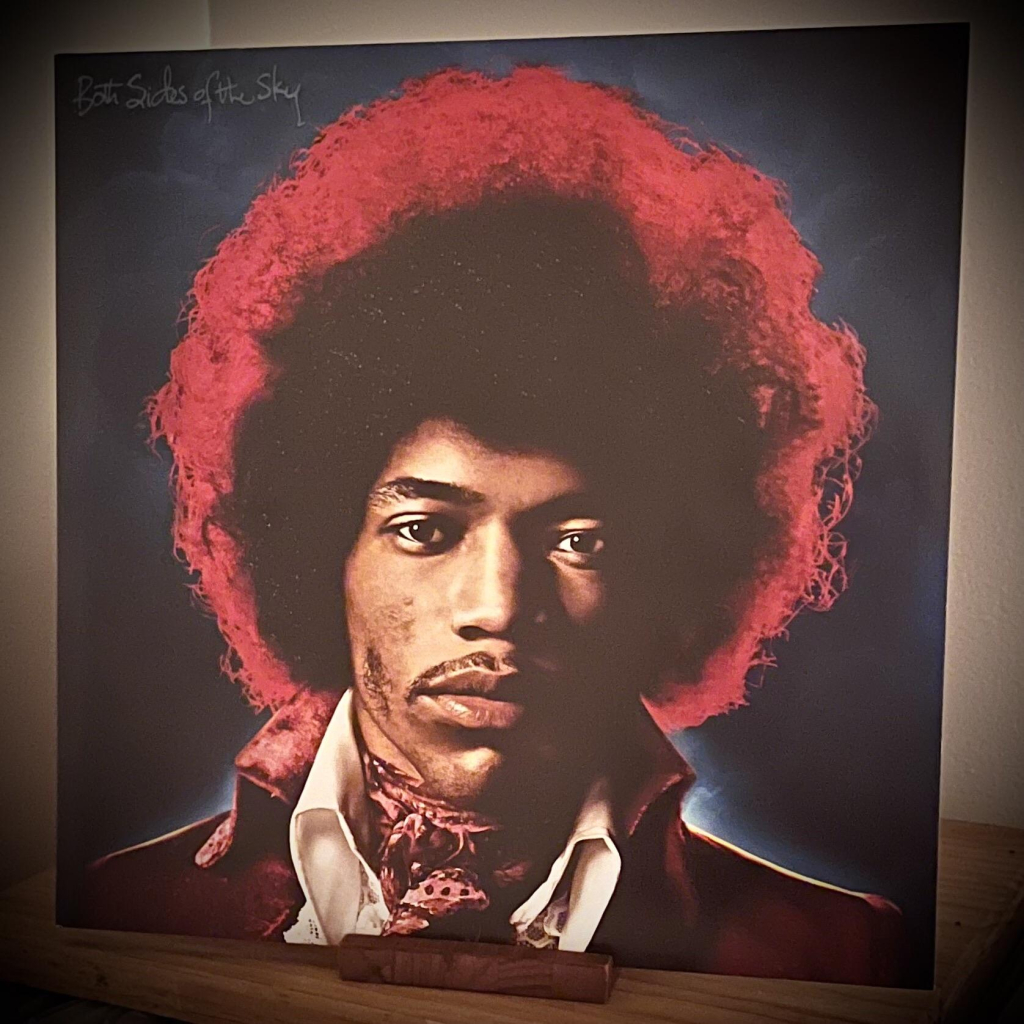 The image features the album cover "Both Sides of the Sky," showcasing a portrait of Jimi Hendrix with a stylish outfit. The background has a gradient of blues and reds, enhancing the vibrant appearance of the subject.