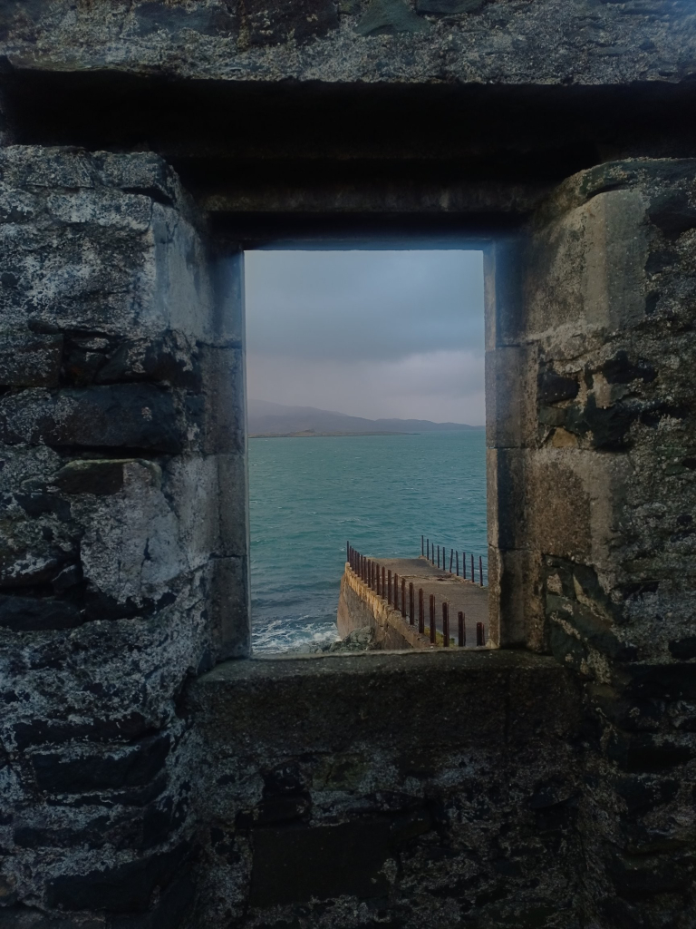 A view through a derelict window, an abandoned and eroding pier just out into the Sound of Jura, the sea mid blue and choppy, varying shades of grey wind blown cloudy sky, islands on the horizon. All bathed in delicate sunset light to the West (left of frame)