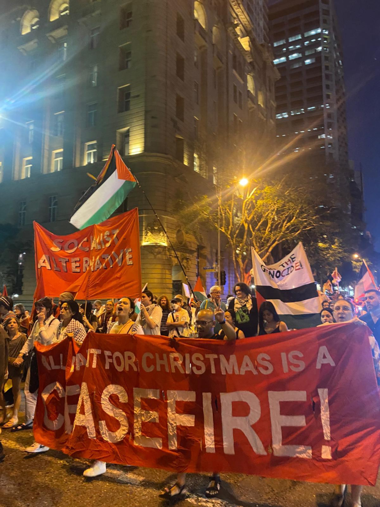 A large crowd of people marching on the street with large banners that read: "all I want for Christmas is a ceasefire", "socialist alternative", "stop the genocide"