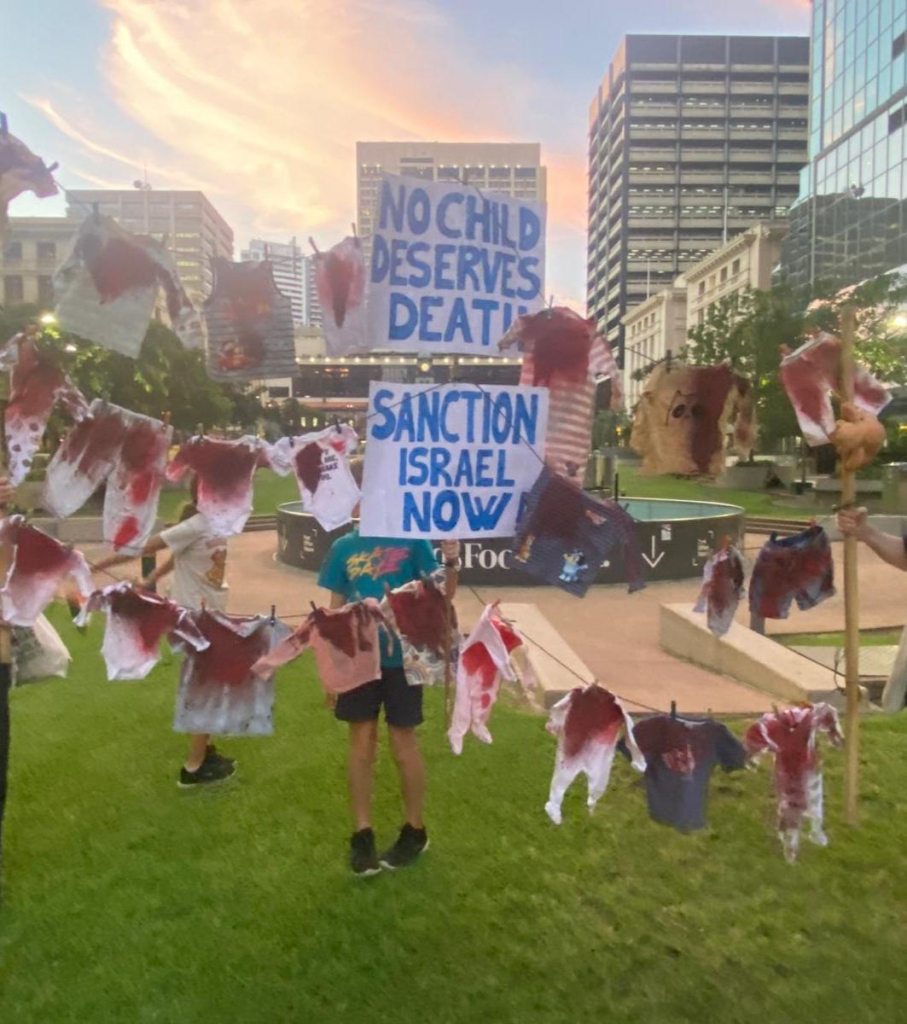 Three lines of children's clothes stained with red paint hung between posts. In the middle, two signs read: "no child deserves death" and "sanction Israel now"