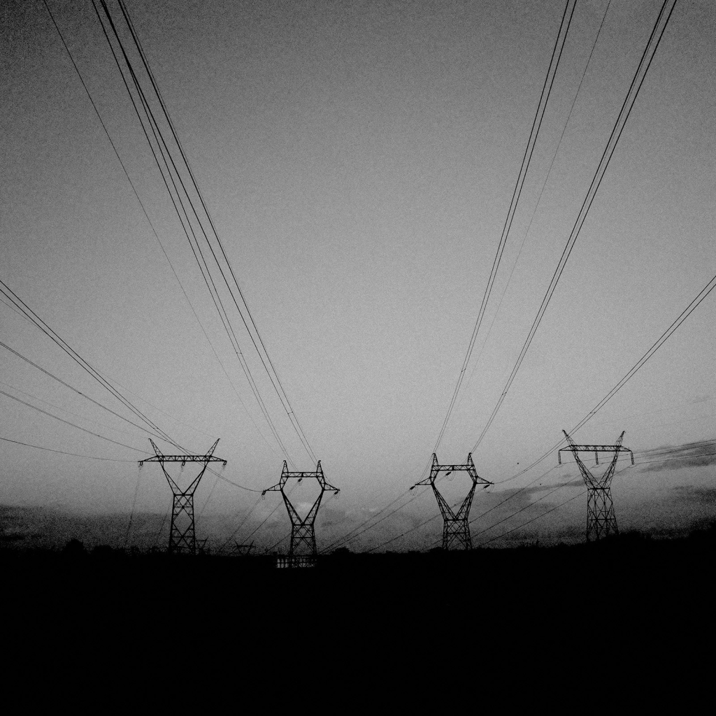 Photo de quatre pylônes électriques installés côte à côte, et dont les câbles passent au dessus du photographe, dessinant des traites dans le ciel.