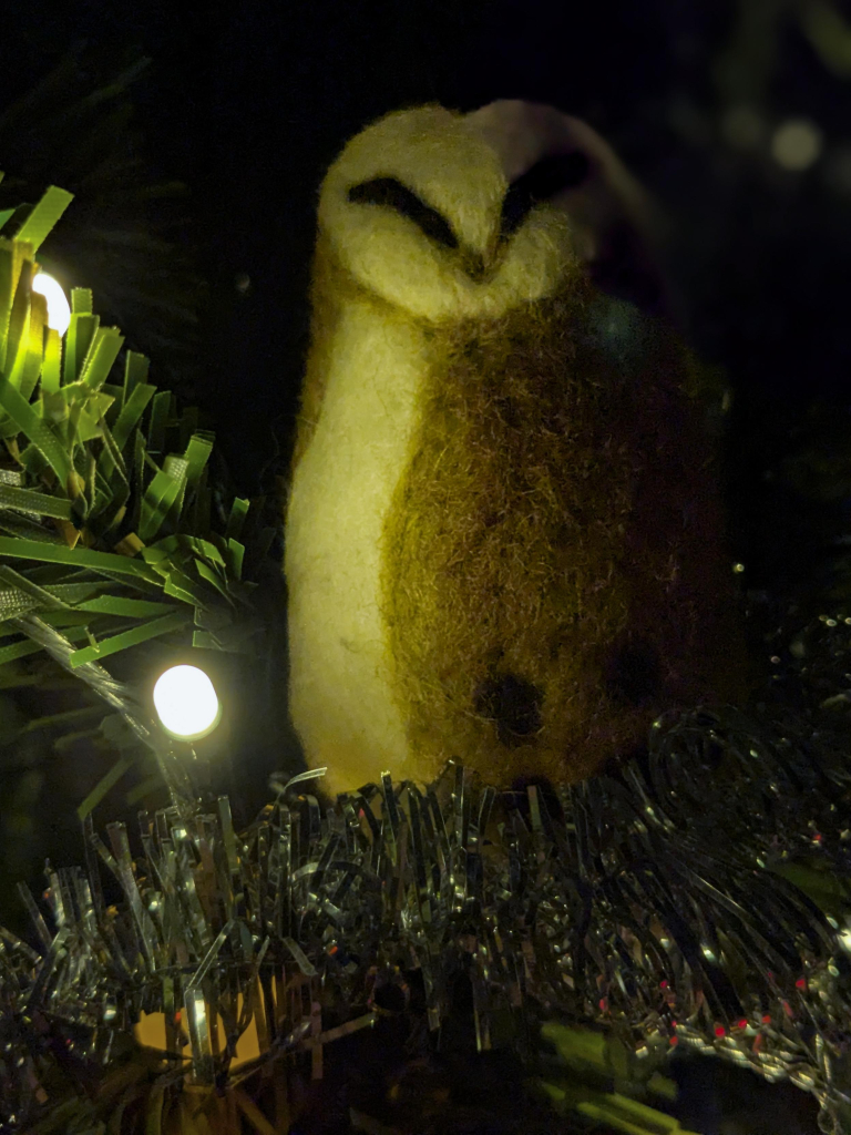 Colour photograph of a felted barn owl decoration nestling in silver tinsel on an artificial Christmas tree. There are also fairy lights.