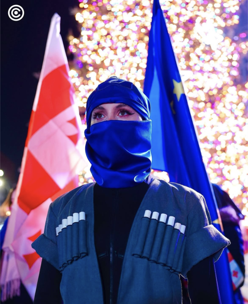 Khorumi Dancer outside parliament in Tbilisi, Georgia. She is wearing a traditional costume and a blue headscarf that shows just her eyes. Behind her are a Georgia flag and an EU flag. Behind the flags are the lights of the Christmas tree.