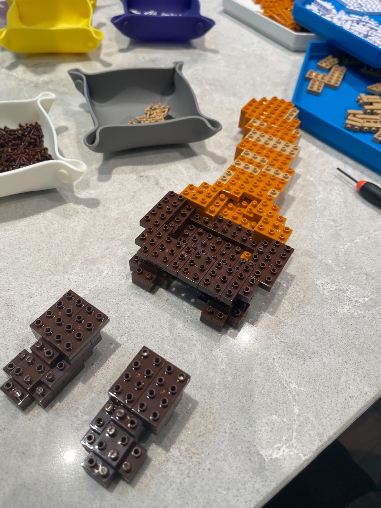 A Jekca figurine, assembly in progress. It’s made from small plastic bricks bolted together with tiny plastic bolts. So far the figure has two dark brown front feet and a stripy orange tail. What could it be??