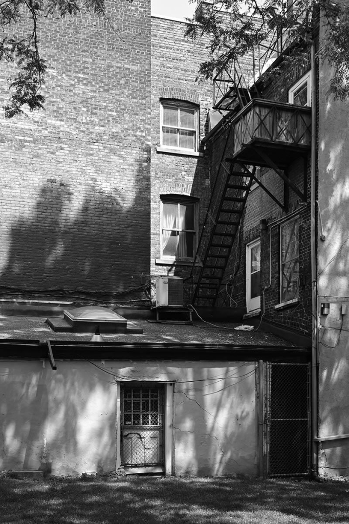 Black and white image, portrait format, rear of an old brownstone building with fire escape...