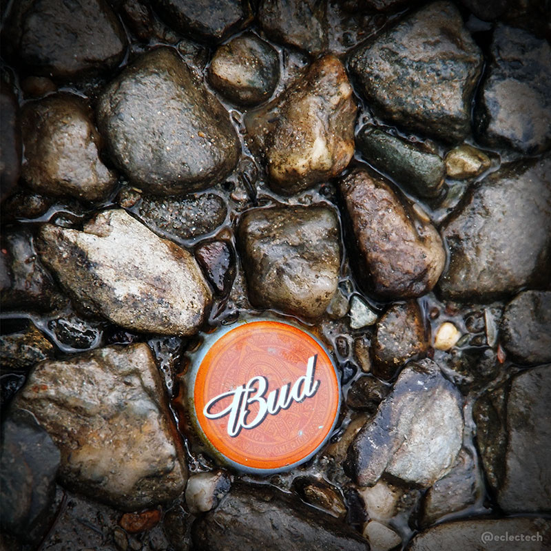 A square photo of a selection of small pebbles set in the muddy edge of a loch. Near the bottom, in the centre, set in the mud surrounde dby the pebbles, is a bright orange Budweiser bottle top with the word Bud written on it in white script. I really dislike littering, but I sort of like this.