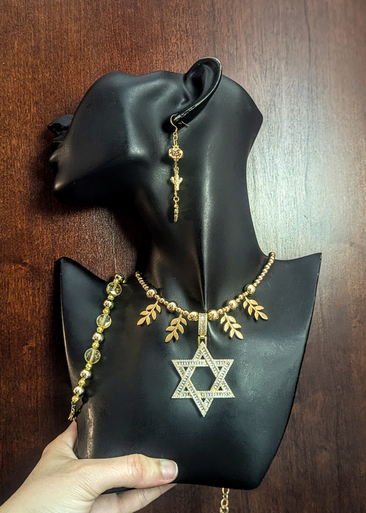 On a black doll, golden Magen David large necklace with golden leaf pendants, a citrine and golden beaded bracelet and drop earrings with pomegranate, cactus and leaf in gold