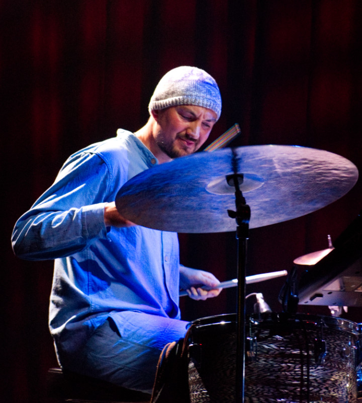 A colour photograph of Corrie Dick, drummer in the duo Norman&Corrie. Wearing a beanie hat and looming over a cymbal, he has a neat beard and moustache. The end of the drumstick in his left hand is just visible over the cymbal; below his left hand holds another stick.