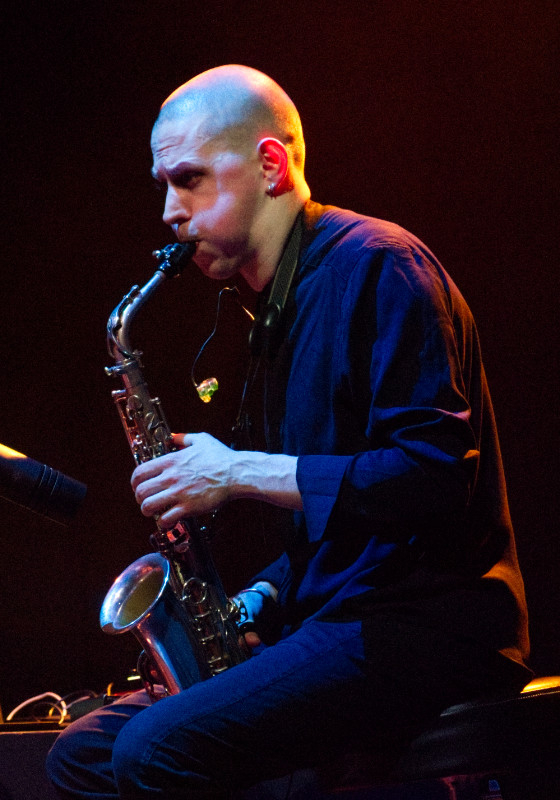 A colour photograph of Norman Willmore,  saxophonist in the duo Norman&Corrie.

His hair is cropped to the skull; he is looking intently at Corrie Dick (not in the photograph) and his cheeks are puffed out as he plays.