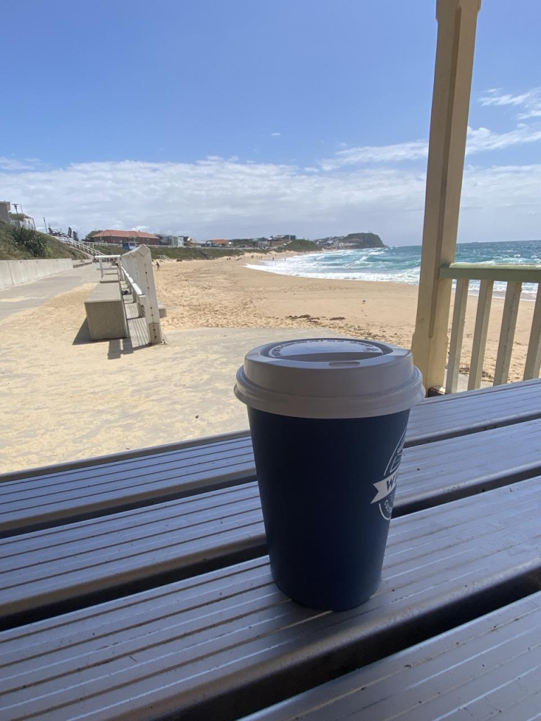 Takeaway coffee cup sitting on a shaded metal picnic table with a view of Merewether Beach looking northwards to Bar Beach directly behind it. Sun shining down on golden sand and blue-green water with lots of white surf breaking along the edge.