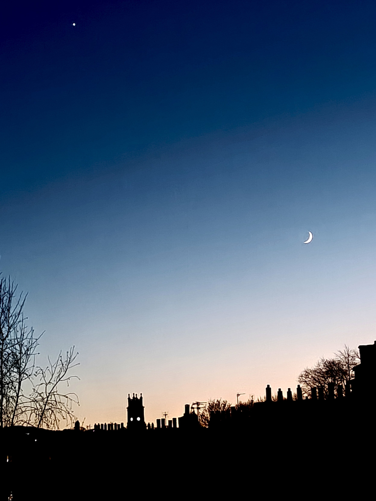 A colour photograph of the early evening sky, which turns from dark inky-blue at the top of the picture through light blue to the orange afterglow of sunset. 

At the bottom of the picture the silhouette of rooftops and chimneys of Canonmills are seen, with the square tower and illuminated clockface of St Stephen's in Stockbridge towards to bottom left of the picture.

On the right of the picture, about halfway up, the slim bit bright crescent of the moon can be seen . Towards the top left is an intense bright dot, which is Venus.