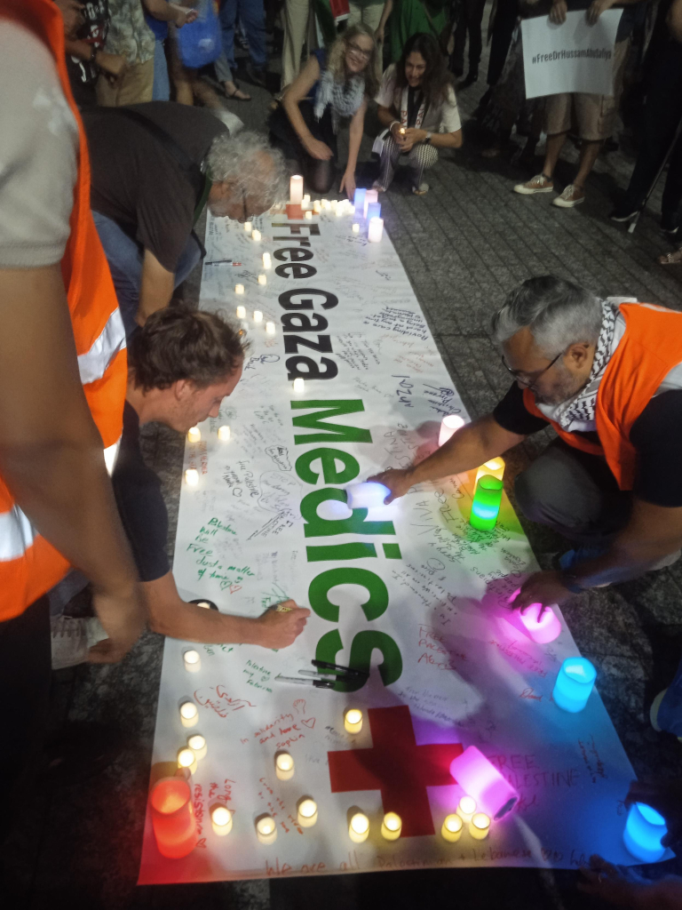 People write their names on a large white banner that reads "free Gaza medics" placed on the ground with candles on top