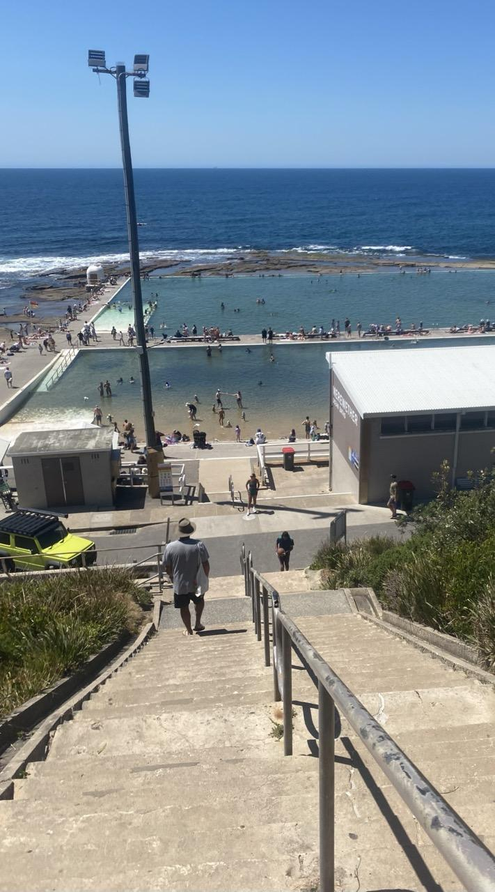 Looking down steep concrete steps that lead to the ocean baths with views out to the ocean horizon. Both pools are visible with water colours that range from light sand or ice green where the water is shallowest, to blue green and even a swatch of navy. It's busy, with many people clustered around the edges. There are a fair number in the pools as well but still plenty of open water. In the closest, shallow pool, it's more families with toddlers in the water. Sunny day with blue skies all the way.