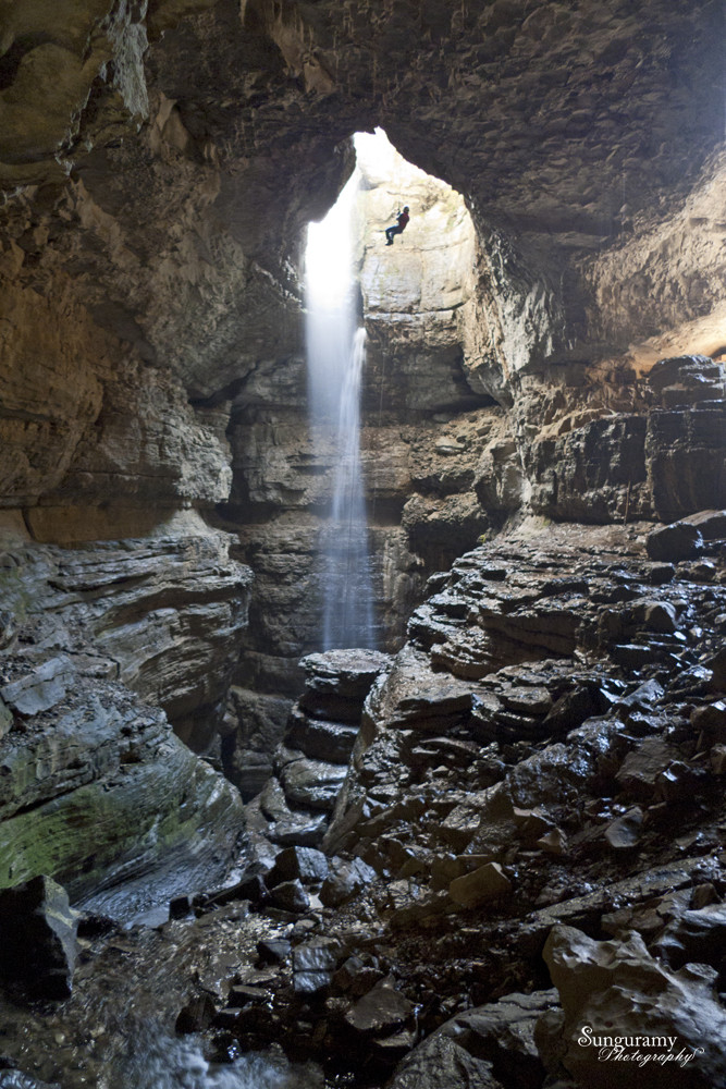 A massive cave chamber with clearly an opening in the ceiling letting in both light, and a massive waterfall. The rock is steep and stepwise, some of it covered in moss. Center lower middle is a very round rock that looks like a nice pedestal. A lone dark figure rappels into the cave, providing scale, near the top where the light is.