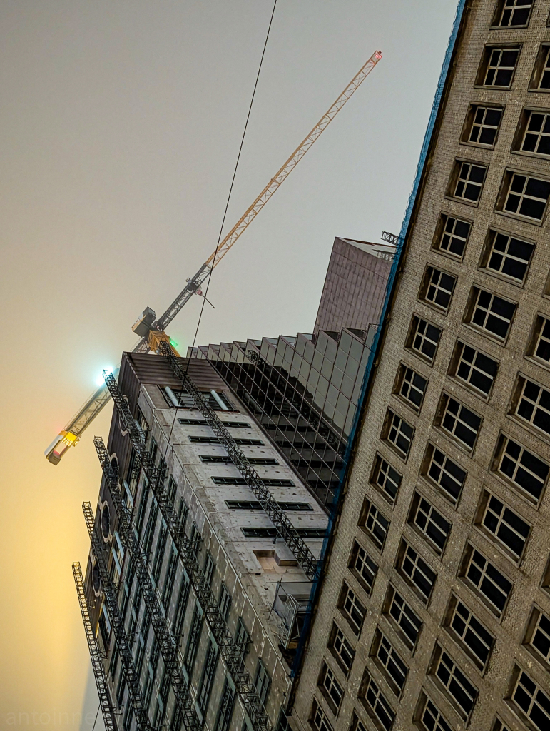 A construction site with tall buildings and a crane against a hazy sky. The building is partially clad in glass and has scaffolding on its facade. The crane is tall and yellow with a long boom reaching upwards. The sky is a muted gray with a hint of orange near the horizon. 