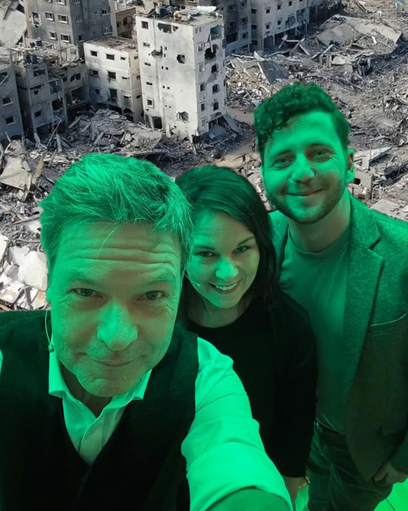 Im Vordergrund ein Selfie von Robert Habeck, Annalena Baerbock und Felix Banaszak in grünem Licht.Im Hintergrund die zerstörte Innenstadt von Beit Lahia, in Gaza.