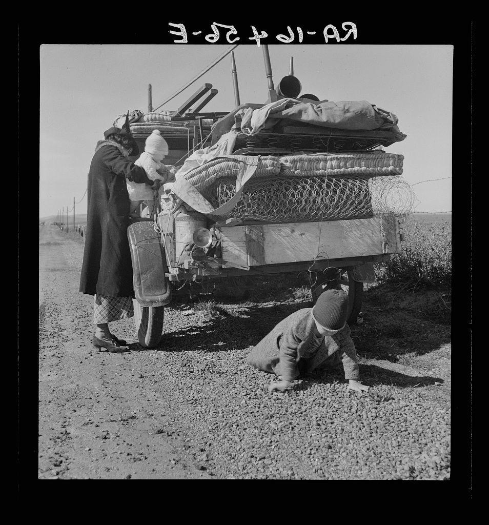 The image depicts a black and white photograph with Hebrew text at the top, indicating it may have been taken in Israel or involving Israeli people. The scene shows three individuals around what appears to be their mode of transportation: an old vehicle loaded down heavily on its bed and back seats.
Two adults are standing beside the vehicle; one is wearing dark clothing while holding a small child wrapped in blankets. They seem focused on something at ground level, possibly attending to luggage or personal belongings strewn across the dusty ground around them. A young boy sits on his knees by the road's edge, seemingly playing with rocks or pebbles.
The surrounding landscape appears barren and dry, suggesting that they may be traveling through a rural area during difficult times, as mentioned in the provided context about Missouri family facing drought conditions. The Hebrew text at the top could provide additional information regarding their journey or circumstances but is not translated here to maintain cultural sensitivity.