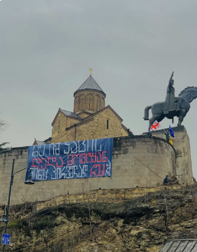 Banner on a wall outside Metheki church, Tbilisi Georgia. The church is on a hill and the banner is on the fortresslike walls next to a statue of a famous Georgian on a horse overlooking the city. There are protestors flying Georgia and EU flags.