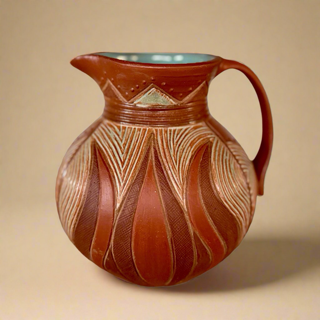 Round pottery pitcher decorated with line carving and geometric patterns. Brick red color exterior, glossy aqua glaze interior. 