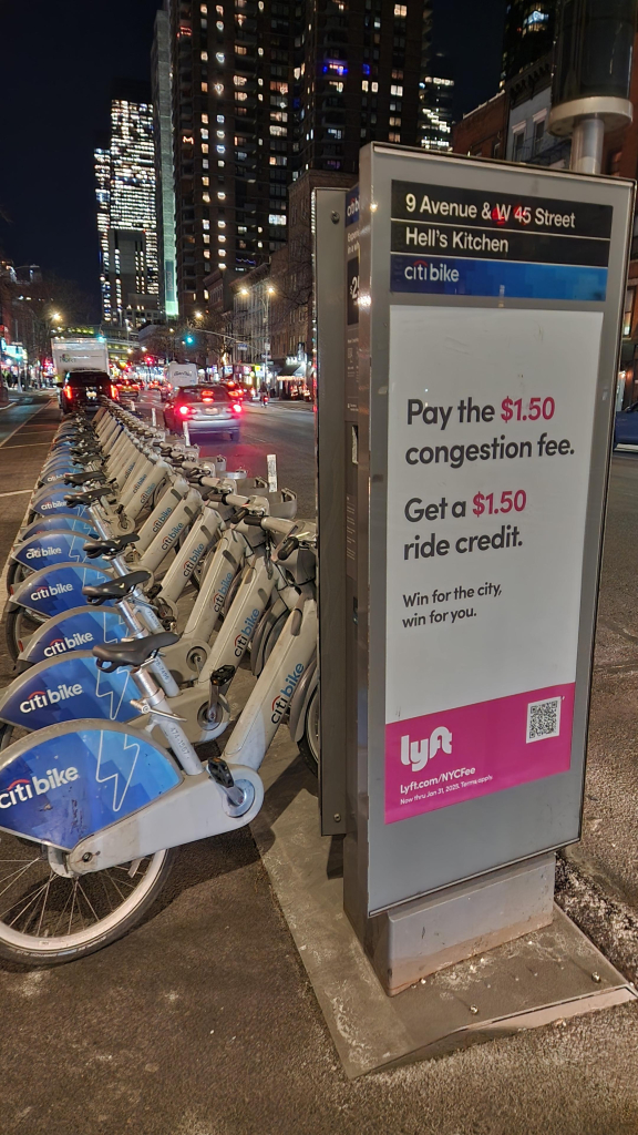 A Citibike station on the left side of Ninth Avenue, housing about two dozen gray ebikes. Apartment and office buildings rise up in the background.

In the foreground there is a rectangular box that is part of the bike station. It houses the payment terminal and has a poster facing traffic. The poster reads:

9 Avenue & W 45 street
Hell's Kitchen
Citibike

Pay the $1.50 congestion fee.

Get a $1.50 ride credit.

Win for the city, win for you.

Lyft