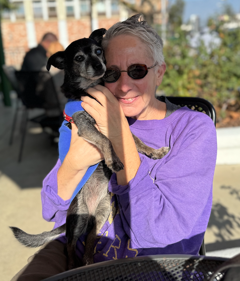 short woman with gray hair, wearing sunglasses and a purple Laker sweatshirt . Sitting outside and holding a gray and black small terrier.