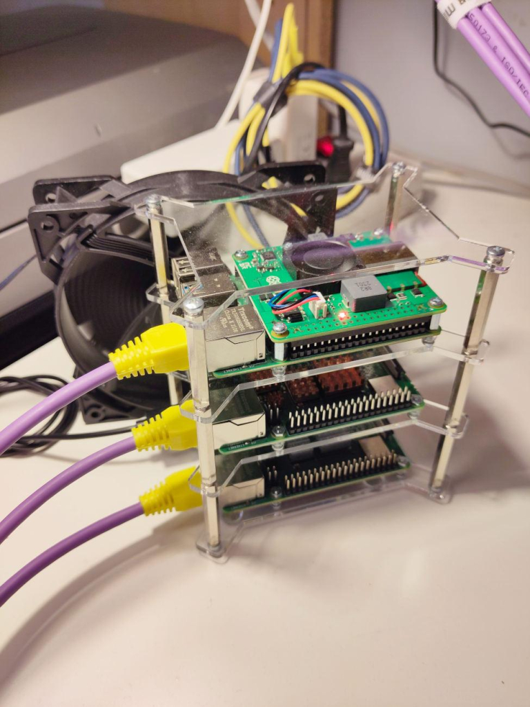 Photo of a cluster of three Rapsberry pis in placed in a single acrylic case holding them one over another. Purple and yellow Ethernet cables com from the Pis at one end. Behind the Pis there is a large black fan blowing air at them. In the background there is a mess of cables powering them.