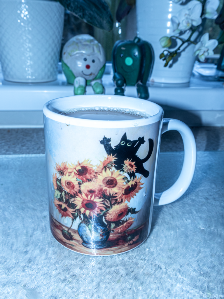 A mug of tea is on the kitchen worktop.  It has a picture of a vase of sunflowers (in Van Gogh style), with a black cat behind, leaping in the air & about to pounce on the flowers.
Behind is the kitchen windowsill with  two pots containing orchids on it, and two little plaster characters based on vegetables (a  cauliflower & a green pepper) sitting on the edge of the windowsill with legs hanging down over the edge.