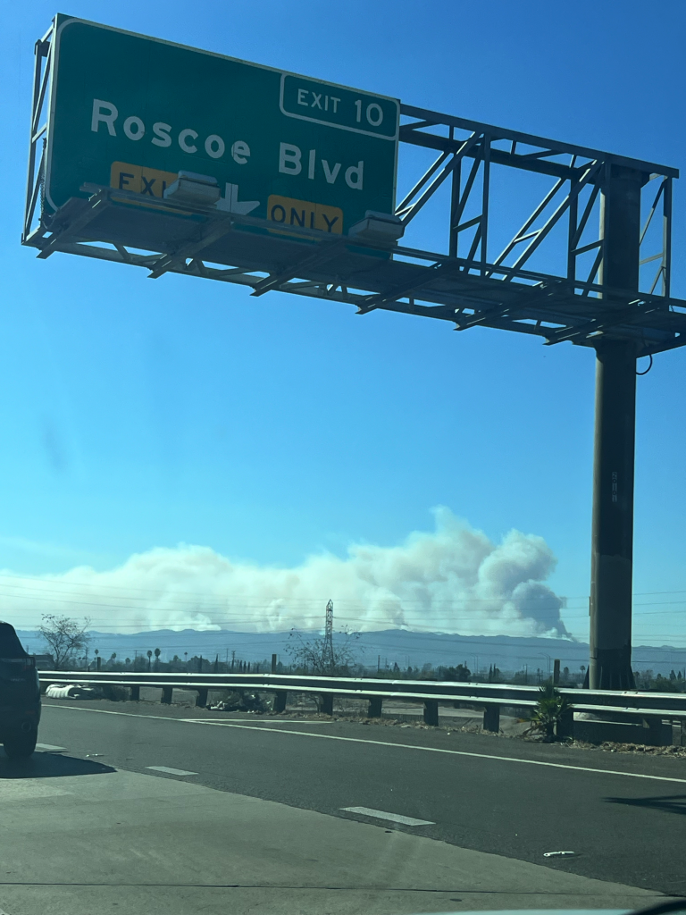 Green exit sign for "Rosco Blvd". Blue sky, and huge smoke plume on the mountain ridge line in the distance. Otherwise, clear blue skies. Taken from the freeway, so we see some road and the metal guardrail