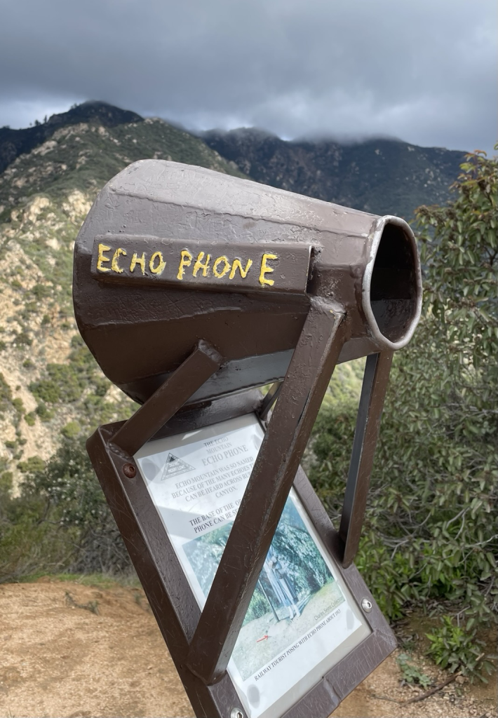 A photo of the Echo Phone with mountainous terrain behind it. The plaque reads, “Echo Mountain is so named because of the many echoes that can be heard across Rubio Canyon. The Mt. Lowe Railway installed an Echo Phone similar to this one in the early 1900s. The base of the original Echo Phone can be seen behind this sign post.”