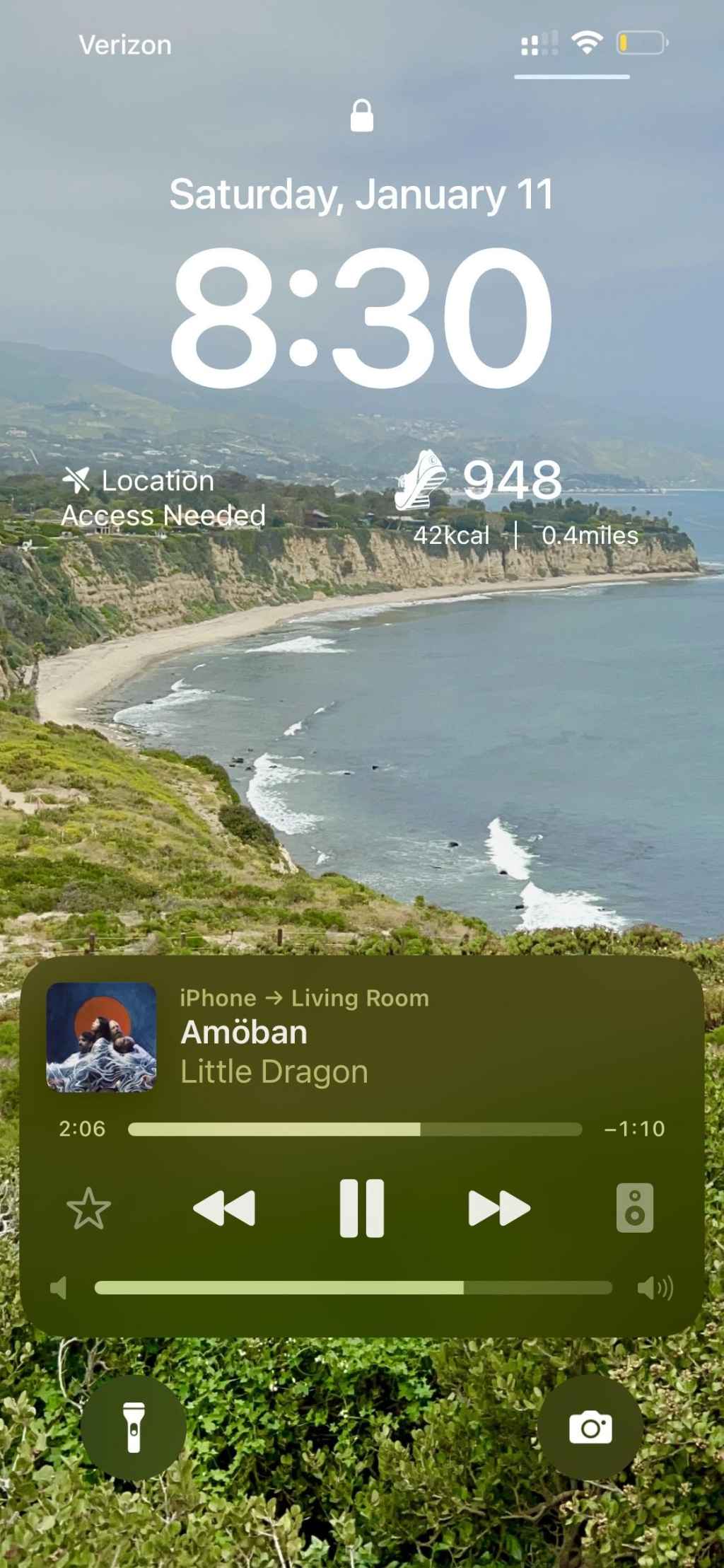 A smartphone screen displaying the time, date with a music player showing the song "Amöban" by Little Dragon. The background features a scenic coastal view with cliffs and a beach. This is at the top of point dume in Malibu, currently at risk in the palisades fire.  